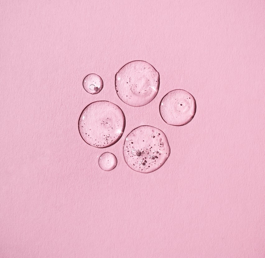 Drops of facial serum or essential oil on pink background.